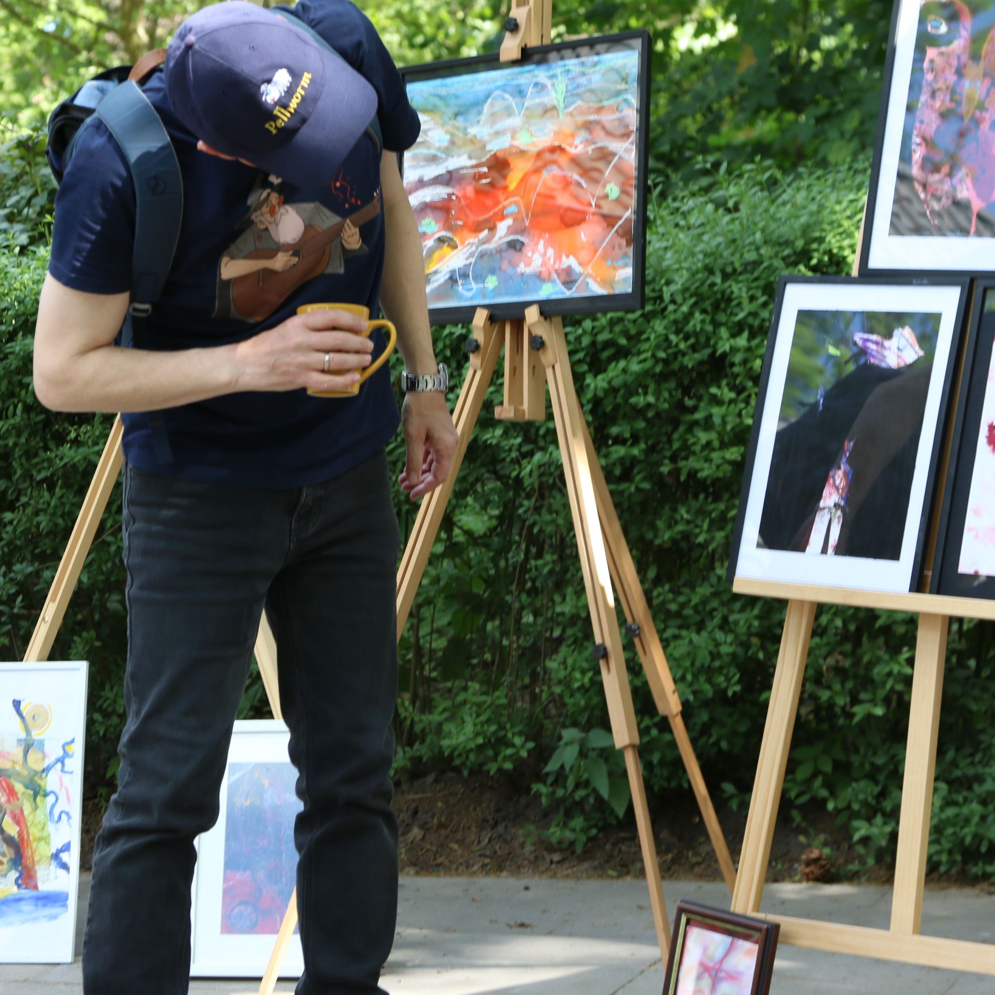 Gemalte Bilder und Fotografien sind draußen im Park ausgestellt. Sie stehen auf Staffeleien und auf dem Boden. Ein Mann bäugt sich herunter, um sich die Bilder genau anzusehen.