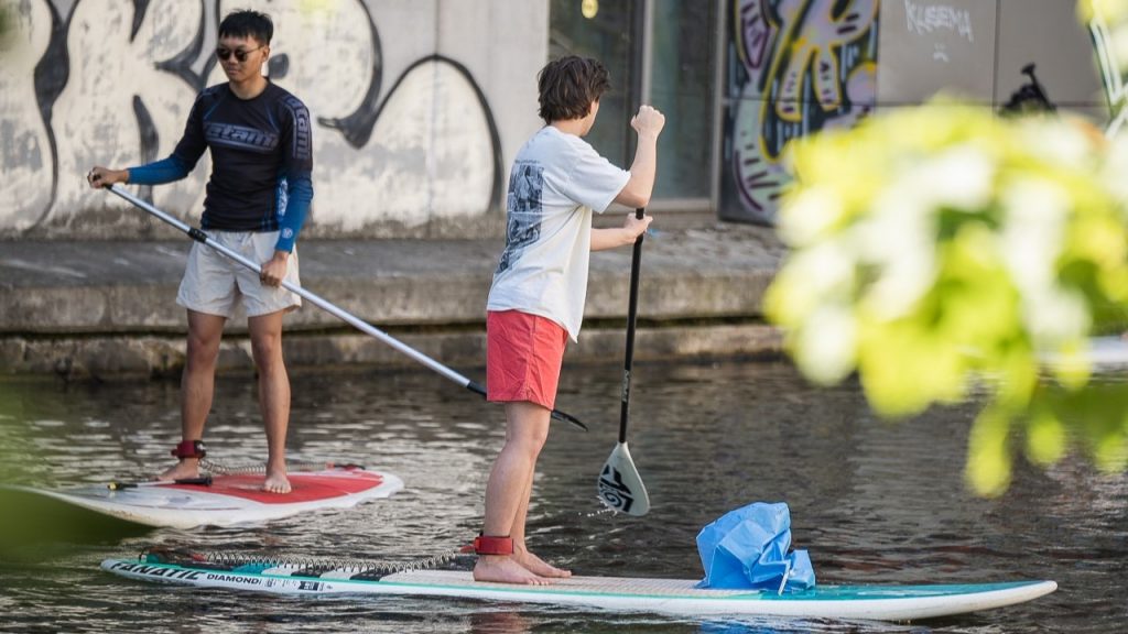 Zwei Männer auf Stand Up Boards paddeln über den Kanal und fischen Müll aus dem Wasser.