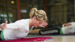 Eine Frau liegt auf dem Bauch mit dem Gesicht zum Boden und hebt ihren Oberkörper nach oben. Dabei reckt sie ihre Arme zur Seite.