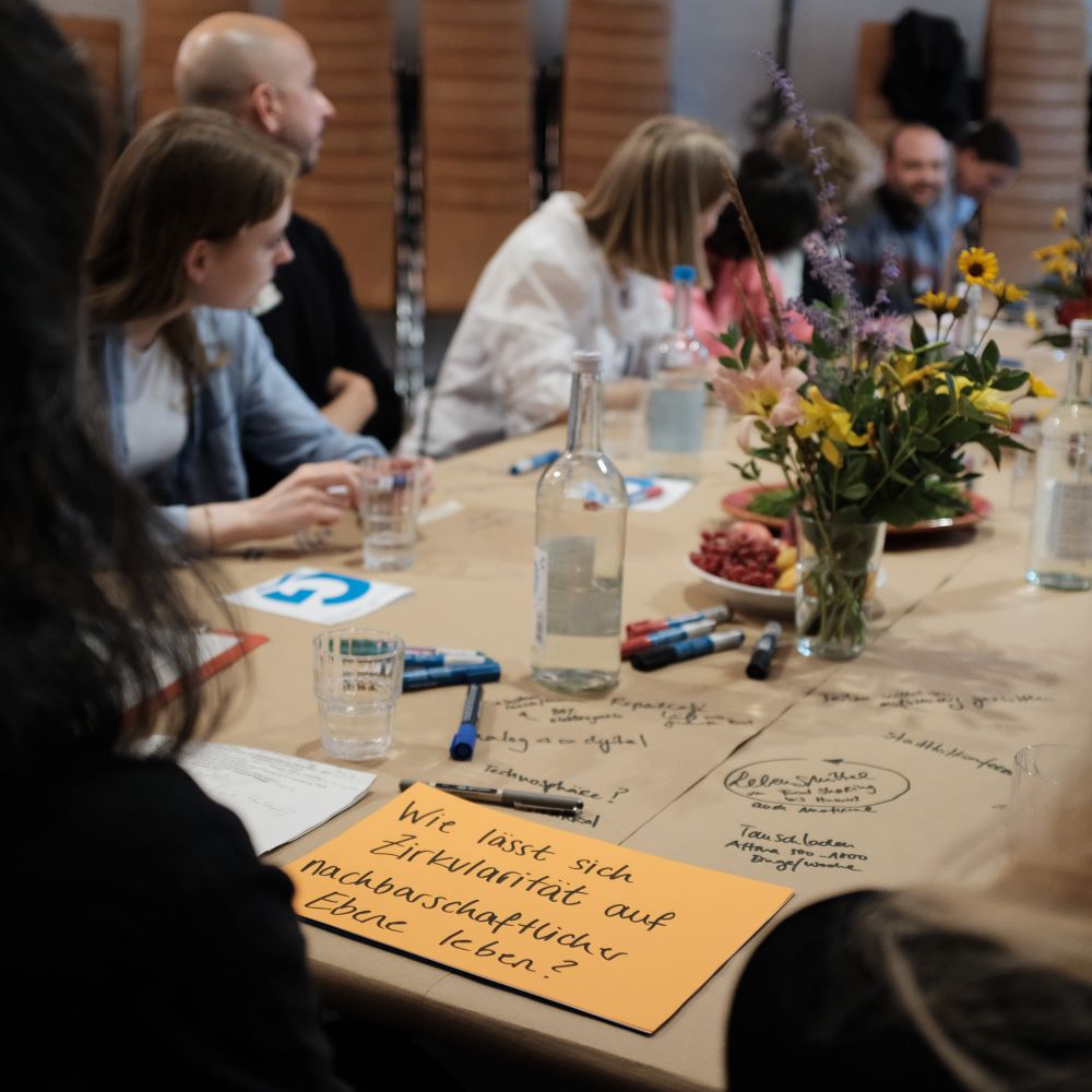 Ein Tisch mit Menschen, die einander zuhören. Im Fokus sind Notizen und Textmarker zu sehen