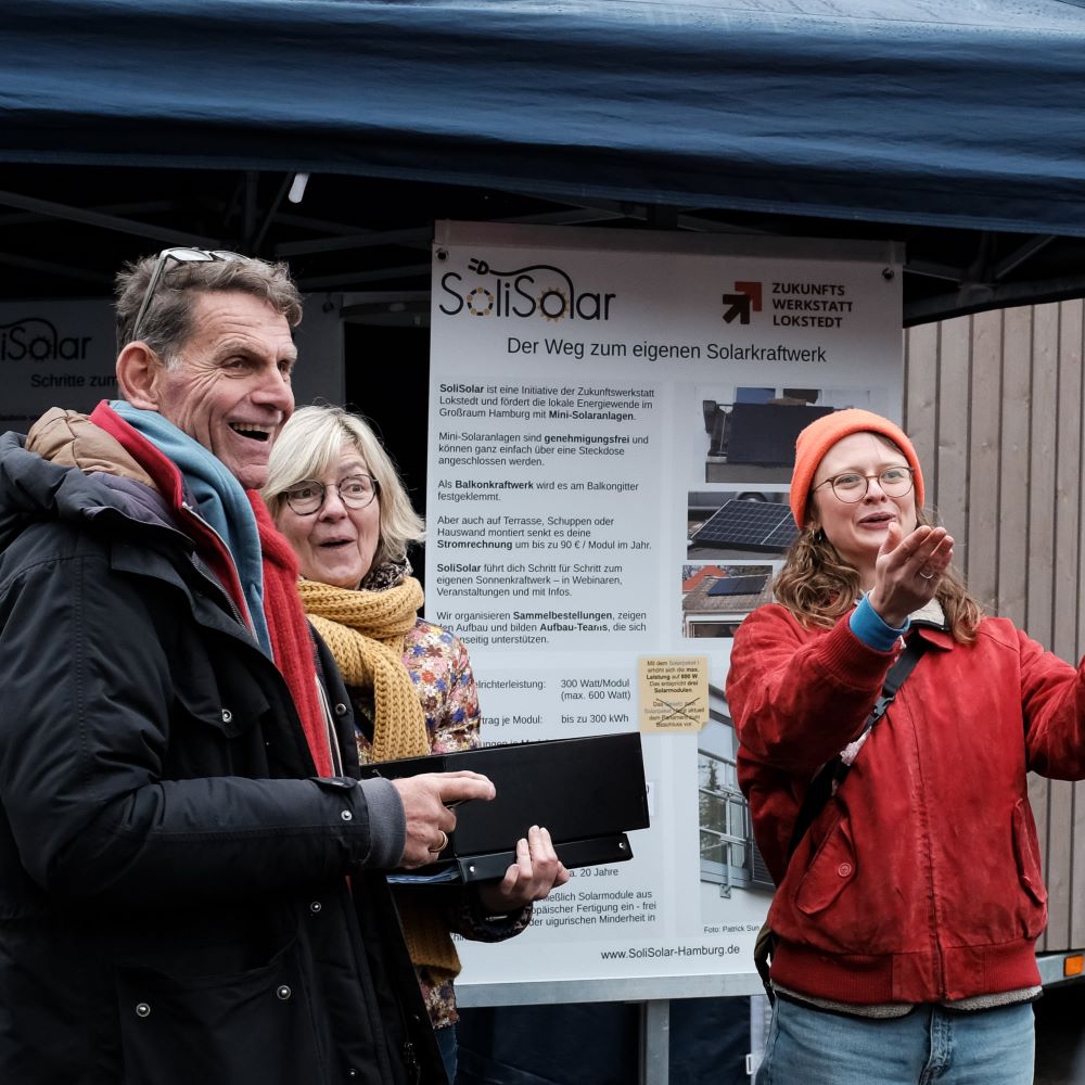 Zwei Frauen und ein Mann lachen. Die eine Frau zeigt auf eine Person im Publikum. Das Bild zeigt die Verlosung des Balkonkraftwerks mit Solisolar.